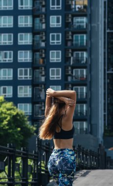 Arka planda, arka planda bina ile kolunu ve omzunu uzatan bir kadın var. Spor kıyafetleriyle şehirde kollarını açmış genç, sıska bir kadının fotoğrafı.