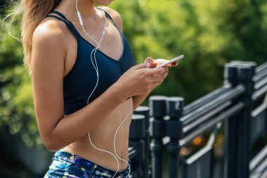 Dışarıda elinde cep telefonu tutan ve kulaklıkla müzik dinleyen tanınmayan fitness kadını .Spor giyim kuşağındaki sağlıklı kadın müzik dinlemek için cep telefonu kullanıyor ve şehirde koşu yaptıktan sonra sağlık durumunu takip ediyor.