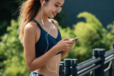Spor giyim bölümünde gülümseyen kadın açık havada müzik dinlemek için cep telefonu kullanıyor. Spor giyim sektöründe formda bir kadın müzik dinlemek için cep telefonu kullanıyor ve şehirde koştuktan sonra uygulamayı takip ediyor.