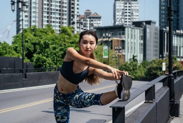 Sporty woman stretching her leg over bridge fence and looking at camera. Confident sporty woman looking at camera and stretching leg on the bridge fence with city view and buildings in background