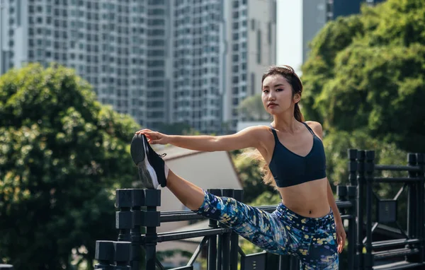 Attractive woman stretching her leg over bridge fence and looking at camera. Confident sporty woman looking at camera and stretching leg on the bridge fence with city view and buildings in background