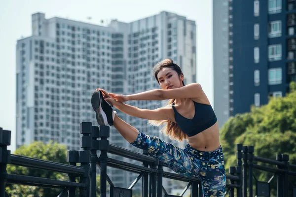 Confident sporty woman stretching leg on the bridge fence and looking at camera. Attractive young woman looking at camera and stretching her leg over bridge fence with city buildings in background