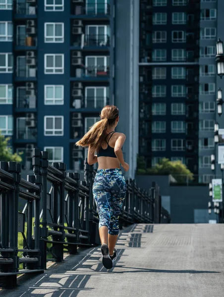 Rear view of woman running on city road with modern buildings in the background. Back view of skinny woman in sportswear jogging in the city
