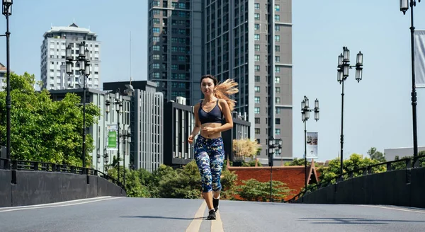 Woman running on city road with buildings in background. Attractive young woman jogging in city with modern buildings in background on sunny morning