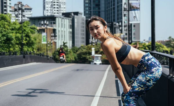 Tired woman taking a break of running and looking at camera at city road with modern buildings in the background. Exhausted jogging woman taking break to breathe