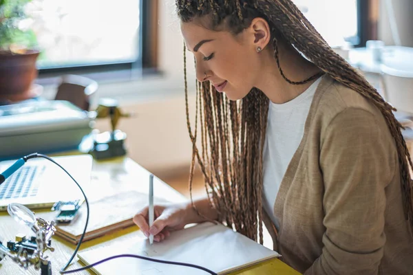 Smiling woman writing at desk. Female student studying electronics engineering, sitting at the table writing notes at class. Studying online, e-learning student, distance learning