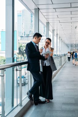 Modern koridorda durup tablete bakan mutlu iş arkadaşları. İş adamı ve iş kadını ofis koridorunda ayakta durup iş stratejisini tartışıyor ve dijital tableti iş için kullanıyor.