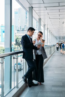 Modern koridorda durup tablete bakan mutlu iş arkadaşları. İş adamı ve iş kadını ofis koridorunda ayakta durup iş stratejisini tartışıyor ve dijital tableti iş için kullanıyor.