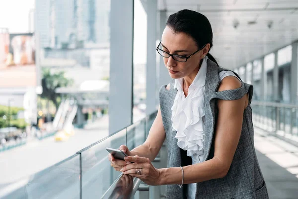 Business woman using mobile phone outside. Business woman talking with business partner online on mobile phone while standing in modern balcony at work