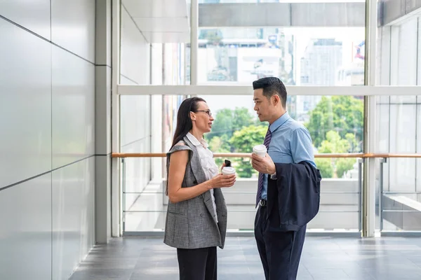 Happy business colleagues standing in modern hallway with big windows, talking and drinking coffee together. Elegant businesswoman and handsome businessman smiling and discuss about new business project