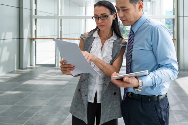 Two cheerful business people reading papers documents and using digital tablet together . Businesswoman showing good news on papers to smiling businessman who is holding white digital tablet