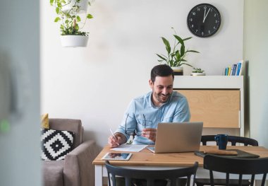 Genç işadamı evde bir masada oturuyor dizüstü bilgisayarla çalışıyor ve kağıda yazıyor. Yakışıklı gülümseyen adam masasında oturuyor, kağıtları dolduruyor, evden çalışıyor, video görüşmesinde konuşuyor.