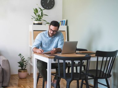 Genç bir iş adamı evde oturmuş, internette dizüstü bilgisayarda çalışıyor ve kağıda yazıyor. Gülümseyen işadamı dizüstü bilgisayarlı ve tabletli masada oturuyor, online toplantı yapıyor, internet konferansı yapıyor.