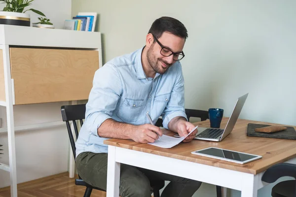 笑顔の男はノートパソコンやタブレットでテーブルに座って 紙に書き込み Web会議を持っています 若いビジネスマンはノートパソコンで自宅から仕事のテーブルに座って紙に書く — ストック写真