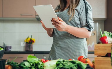 Modern mutfakta yemek pişirirken dijital tabletten yemek tarifi okuyan tanınmayan bir kadın. Beyaz tabletle sağlıklı yemek yapmak için internet eğitimi izleyen ev kadınının kesilmiş fotoğrafı.