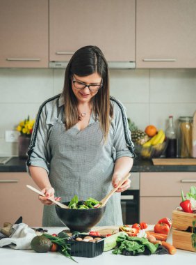 Güzel bir kadın evde taze salata yapıyor. Neşeli ev hanımı öğle yemeği hazırlıyor, organik sebzeleri mutfak masasında kaşıkla karıştırıyor, sağlıklı beslenme konsepti.