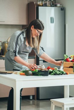 Modern mutfakta yemek pişirirken dijital tabletten yemek tarifi okuyan neşeli bir kadın. Mutlu ev hanımı internetten sağlıklı yemek yapma dersleri izliyor beyaz tablet üzerinde, taze organik sebzelerle dolu bir masanın önünde duruyor.