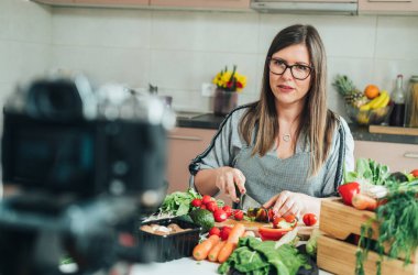Güzel bir kadın, çevrimiçi yemek kursunun video dersini ev mutfağında kaydediyor. Kadın aşçı mutfak masasında taze sebze kesiyor ve YouTube yemek kanalı için video çekiyor.