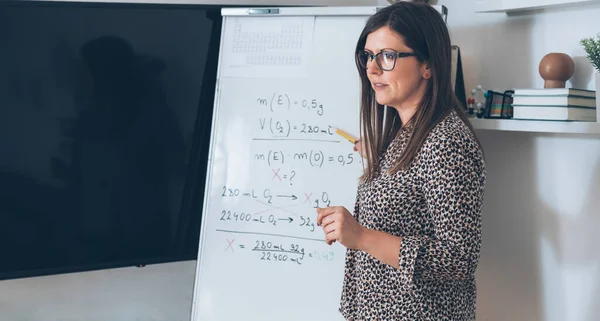 Teacher recording video tutorial for e-learning classFemale professor standing and explaining the assignment pointing on the whiteboard and using camera for live streaming online on the internet for Physic students, remote education class.