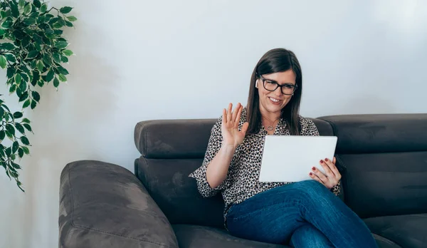 Smiling Businesswoman Say Hello to Teamwork Colleague in Video Conference Meeting while Sitting on Couch at Home.Happy business woman with glasses looking at digital tablet and waving with hands, welcoming clients or coworkers at web conference.