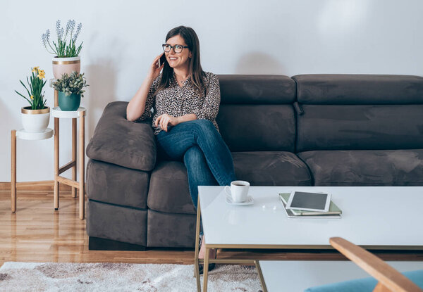 Счастливая деловая женщина сидит на диване и разговаривает на мобильном телефоне. Веселая улыбающаяся деловая женщина отдыхает на диване в гостиной дома и звонит кому-то на смартфон.