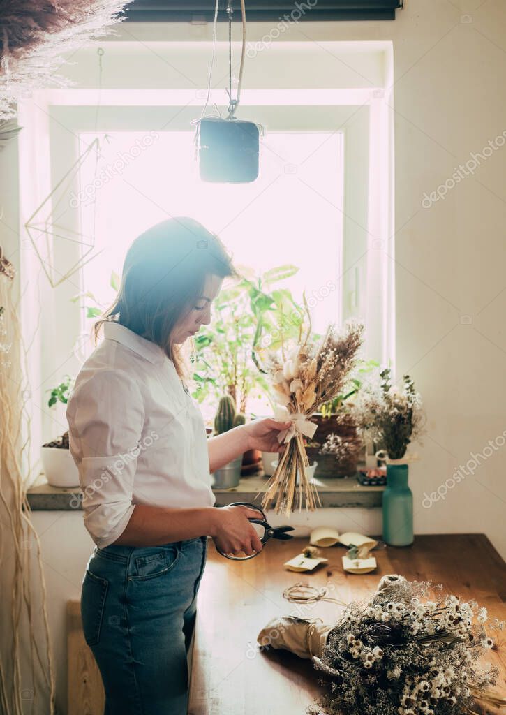 Beautiful Florist Arranging Bouquet of a Dry Flowers at her WorkshopYoung talented woman cutting with scissors dried plants and flowers arrangement in her flower shop, sustainable lifestyle concept.