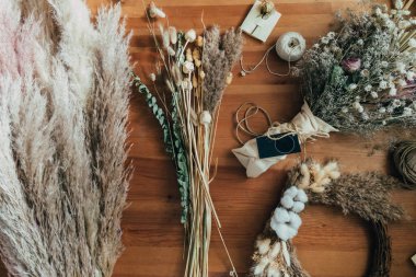 Kurumuş Çiçekler ve Bitkiler Masası, Everlasting Bouquet, Tahta Masa 'da Çelenk ve Pampas Çimeni. Küçük işletme atölyesindeki çeşitli kuru çiçekler, kır çiçekleri, otlar, çelenkler, ipler ve hediye kartlarının fotoğraflarının hemen üstünde..