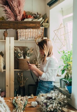 Güzel Bayan Çiçekçi, atölyesinde Tamamen Farklı Kuru Çiçekler ve Bitkilerle dolu cep telefonu kullanıyor. Sanat ve zanaat stüdyosunda duran ve akıllı telefonuyla müşterilerine ve müşterilerine mesaj yazan ya da e-posta gönderen yetenekli genç bir kadın..