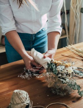 Çiçek dükkanındaki Wooden Masası 'nda kurumuş çiçek buketini saran Bayan Çiçekçinin Elleri. Tanınmayan bir kadın girişimcinin atölyesinde bir buket kuru çiçek ve bitkiler düzenlemesi ve çalışma masasının üzerinde fotokopi alanı olması.