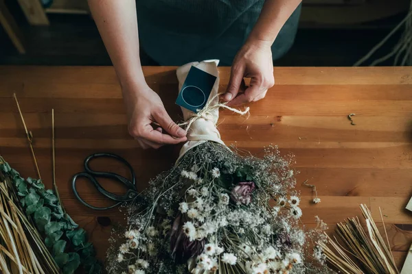 女性の花屋の手彼女の花店で木製のテーブルでドライフラワーの永遠の花束を作る 認識できない女性起業家の高い角度ビューは 机の上にコピースペースと彼女のワークショップスタジオで乾燥した花や植物の花束を手配 — ストック写真