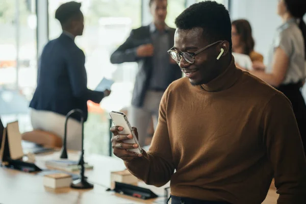 Smiling Business Man Using Mobile Phone and Bluetooth Earphones at Work.Cheerful African-American businessman enjoying listening to music on wireless earphones and smartphone while leaning on the table in open plan office with multi-ethnic team.