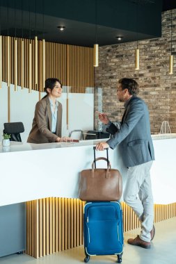 Kadın otel resepsiyonisti, iş adamına valizli kontrol için yardım ediyor. Otel resepsiyonunda kapıcı ile hapşırık koruması ile konuşuyor..