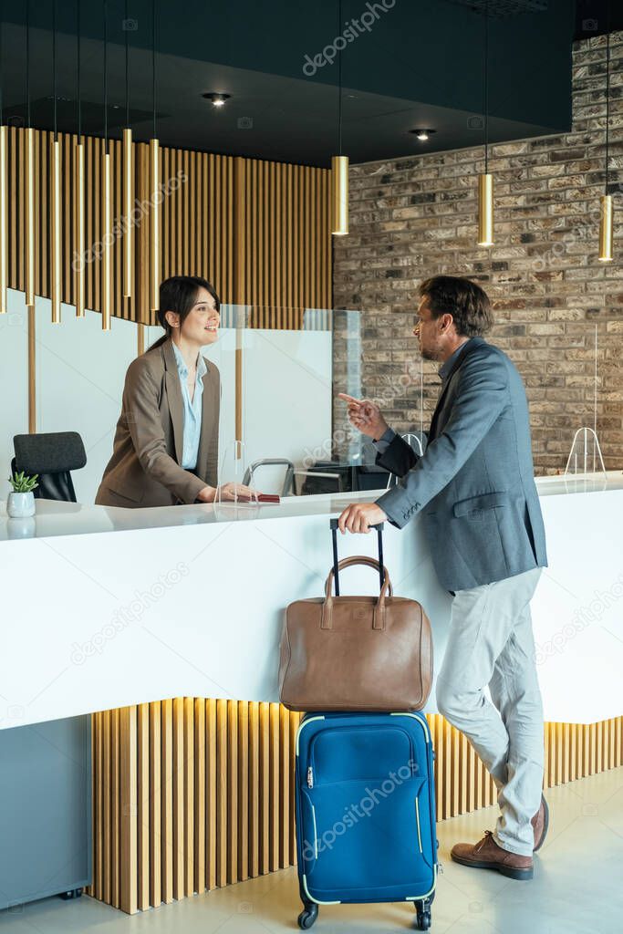 Female Hotel Receptionist Assisting Businessman for Checking In.Business man with luggage talking with concierge on hotel reception with sneeze guard protection.