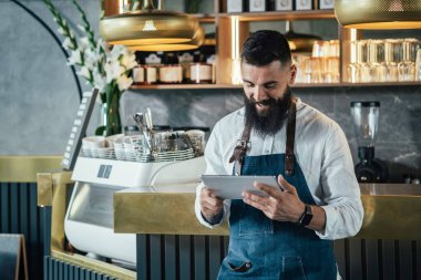 Bir kafede dijital tablet kullanan yakışıklı bir barista. Bar tezgahına yaslanmış, internetten sipariş veren ya da restoranda çalışırken dijital tabletten bir şeyler izleyen gülümseyen garson..