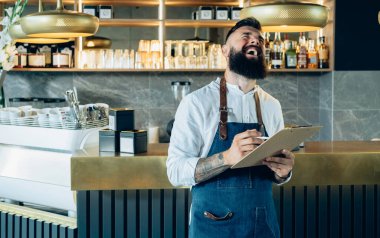 Neşeli Gülümseyen Barista bir kafede ürünlerin envanterini çıkarıyor. Sakallı, gülen, bar tezgahında duran ve restoranda hisse senedi saymak için pano kullanan mutlu garson..