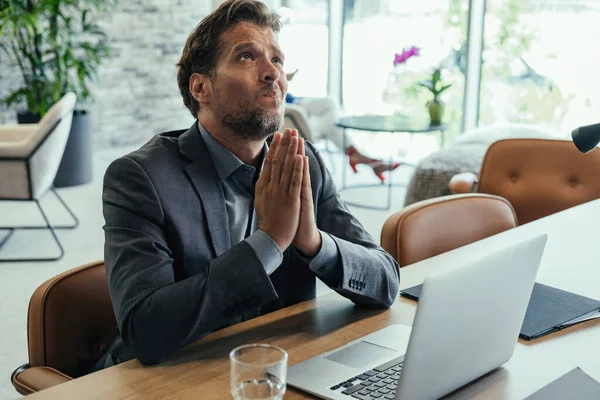 Worried Business Man Praying at Work. Stressed businessman expecting results and praying while sitting at desk and working on laptop computer in the office.