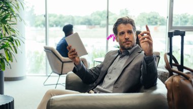 İş adamı, İş Salonunda Uçuşunu beklerken Dijital Tablet kullanıyor. Koltuğunda oturan ve havaalanı lobisinde bekleyen eli havada yakışıklı bir iş adamı..