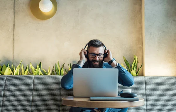 Handsome Businessman Putting Wireless Headphones Before Online Meeting in a Cafe. Cheerful smiling business man with headphones having conference call meeting online on laptop computer at restaurant.