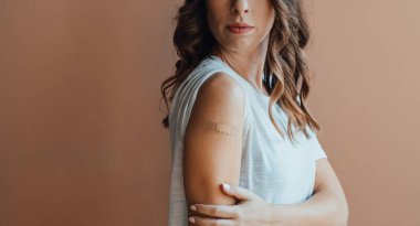 Aşı sonrası koluna bandaj süren isimsiz genç bir kadın. Ciddi bir kız, Covid-19 aşısını aldıktan sonra pembe arka planda dururken kolunu alçıyla gösteriyor..