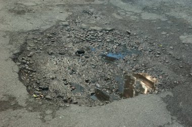 Çakıllarla dolu bir çukur ve binayı yansıtan bir su birikintisi olan kırık asfalt..