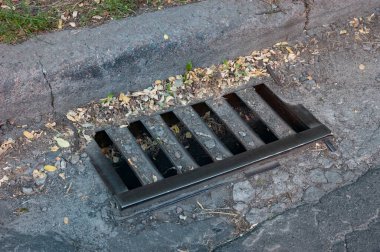 Kaldırım kenarında eski metal rögar ızgarası ve yakında düşen küçük bir sonbahar yaprağı. Mevsimsel dokunmatik ve yıpranmış yüzey dokusuna sahip kentsel ayrıntı.