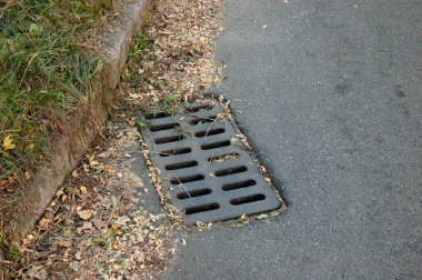 Kaldırım kenarında eski metal rögar ızgarası ve yakında düşen küçük bir sonbahar yaprağı. Mevsimsel dokunmatik ve yıpranmış yüzey dokusuna sahip kentsel ayrıntı.