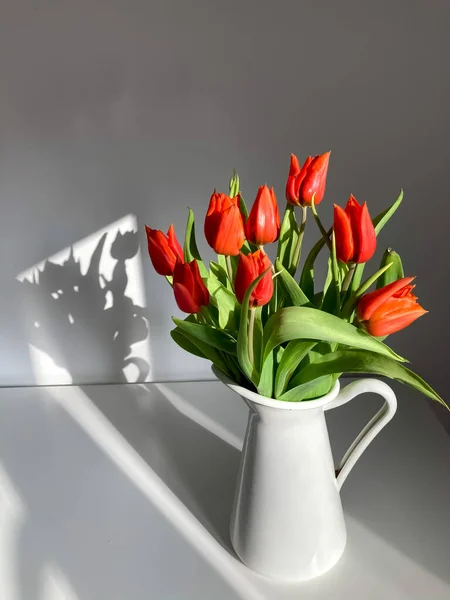 Beyaz bir zemin üzerinde beyaz bir sürahi içinde kırmızı laleler buketi. 