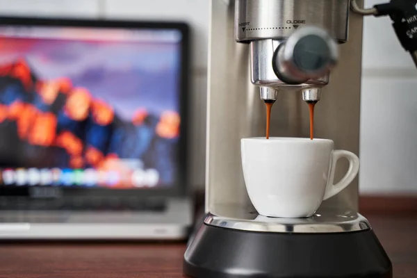 Espresso bir kahve fincanına akar. Bulanık arkaplanda görünür bir bilgisayar var. Merkez ofis ve şirket sahneleri için kullanılabilir.