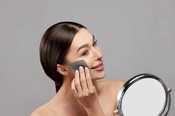 Skin care. Young woman removing oil from face using blotting papers looking in mirror. Beautiful girl model with smooth and healthy skin. Beauty concept. Cosmetology, cosmetics. Facial treatment