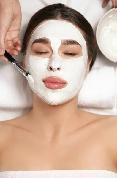 美容師はスパサロンで若い美しい女性の顔に顔マスクを適用する — ストック写真