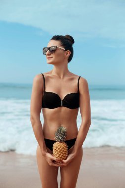 Bikinili bronz tenli güzel kadın ananasları elinde tutuyor.