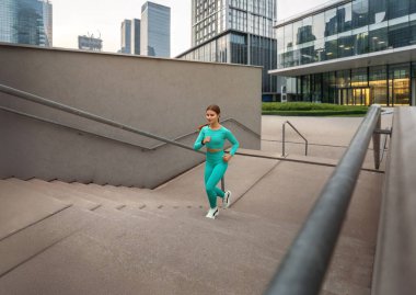 Turkuaz giyimli formalı kadın, şehir ortamında atletizm ve fitness bağlılığını göstermek için açık havada merdivenlerden koşuyor. Egzersiz konsepti