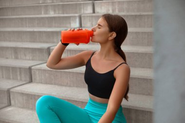 Aktif bayan sporcu, açık hava basamaklarında mola verir, parlak bir içecek konteynırından yudumlayarak, zindelik ve sağlığa olan bağlılığını vurgular. Egzersiz konsepti