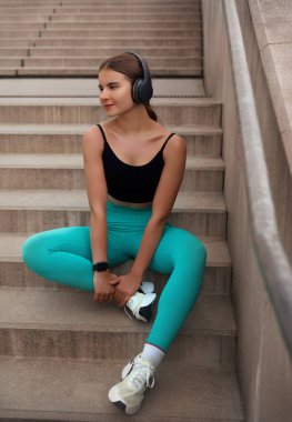 Siyah bluz giyen ve turkuaz tayt giyen sağlıklı bir kadın merdivenlerde oturur, kulaklıkla müzik dinler, sağlıklı bir yaşam tarzı ve uygunluğu somutlaştırır. Egzersiz konsepti